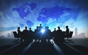 A shadow of a Conference table full of men and women discussing International Business. Projected behind them is a blue map of the world with money signs and a sky scrapper. 