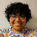 Hispanic female with black curly hair and glasses wearing a colorfully designed shirt.